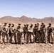 A group of approximately 13 military personnel in desert camouflage uniforms pose for a group photograph in an arid desert environment. They are armed with rifles and wearing patrol caps, standing on rocky terrain with barren mountains visible in the background. The setting and uniforms are consistent with a U.S. Marine Corps training exercise or operational deployment in a desert region.