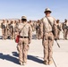 A group of military personnel in desert camouflage uniforms are assembled in formation on a paved surface in an arid desert environment. Two individuals viewed from behind appear to be in leadership or supervisory roles, facing the assembled group. The setting suggests a training exercise, briefing, or formation review at a desert military installation.