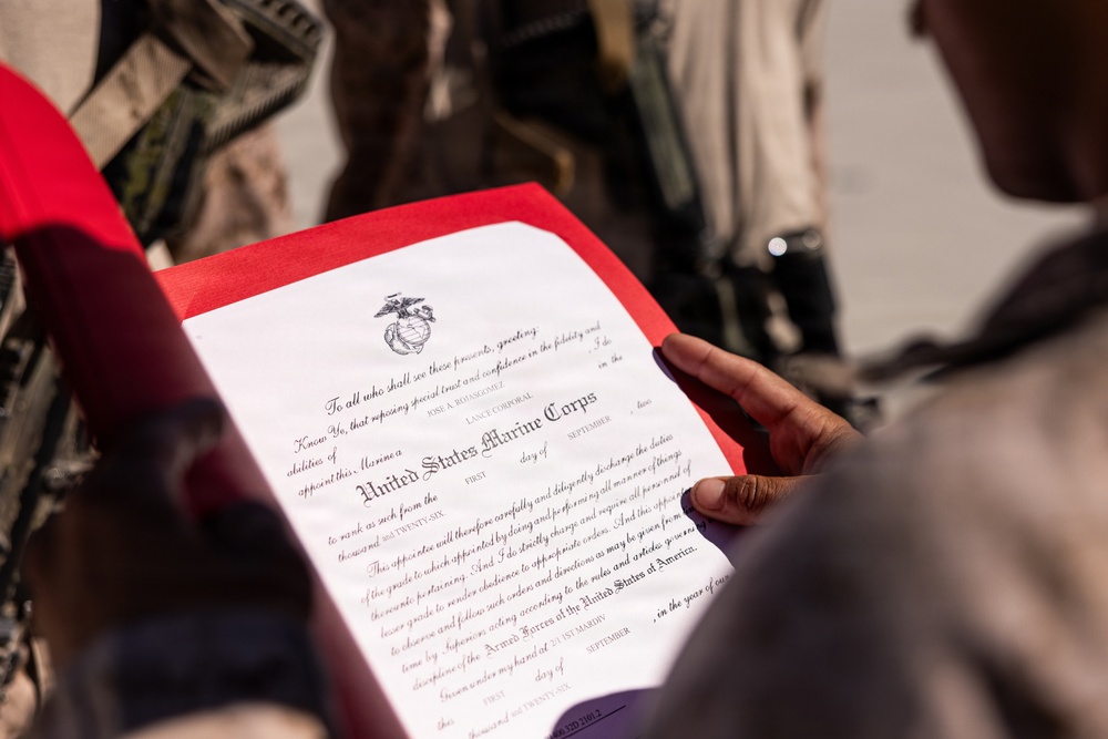 A person is holding an official United States Marine Corps promotion or appointment certificate in a red folder. The document bears the USMC Eagle, Globe, and Anchor emblem and appears to be a Lance Corporal appointment certificate for an individual named Jose A. Bolasegomez. The setting appears to be an outdoor military environment.