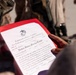 A person is holding an official United States Marine Corps promotion or appointment certificate in a red folder. The document bears the USMC Eagle, Globe, and Anchor emblem and appears to be a Lance Corporal appointment certificate for an individual named Jose A. Bolasegomez. The setting appears to be an outdoor military environment.