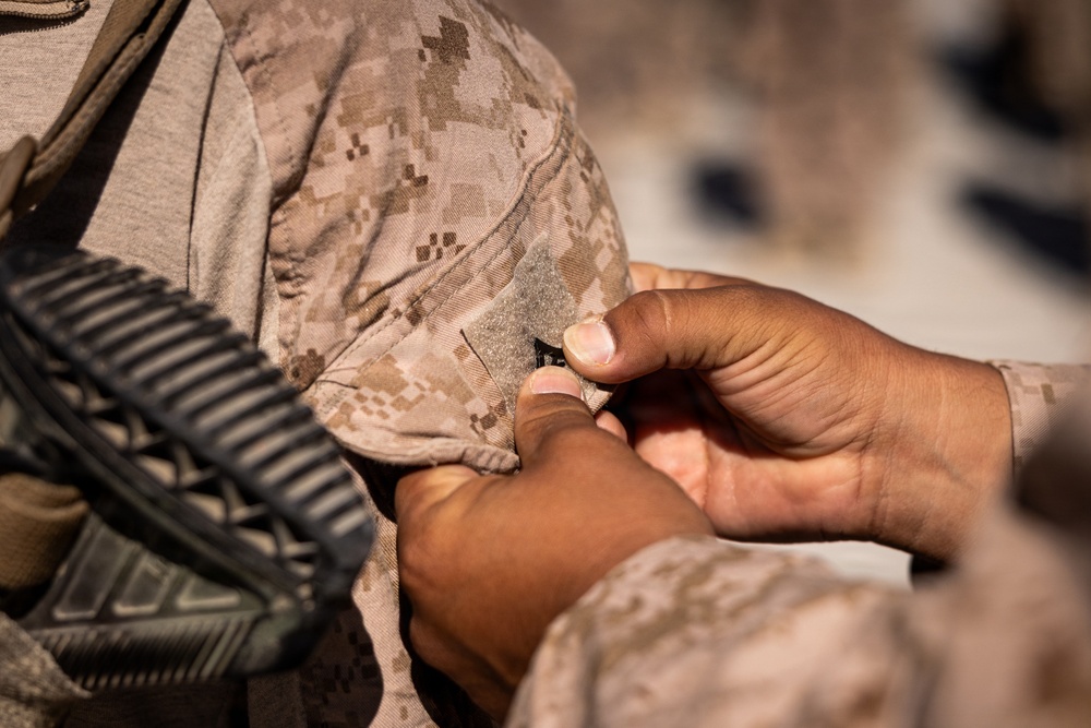 A close-up photograph showing hands manipulating what appears to be a small black item, likely a pin or insignia, on the shoulder area of a desert camouflage military uniform. The uniform pattern is consistent with USMC MARPAT desert camouflage. A black rifle or weapon sling is partially visible on the left side of the frame.