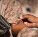 A close-up photograph showing hands manipulating what appears to be a small black item, likely a pin or insignia, on the shoulder area of a desert camouflage military uniform. The uniform pattern is consistent with USMC MARPAT desert camouflage. A black rifle or weapon sling is partially visible on the left side of the frame.
