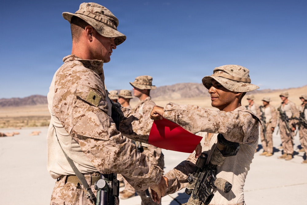 Two military personnel in desert camouflage uniforms are engaged in what appears to be a formal exchange or presentation involving a red item, likely a flag or pennant. The setting is an arid desert environment with mountains visible in the background. Additional uniformed personnel are standing in formation in the background.