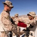 Two military personnel in desert camouflage uniforms are engaged in what appears to be a formal exchange or presentation involving a red item, likely a flag or pennant. The setting is an arid desert environment with mountains visible in the background. Additional uniformed personnel are standing in formation in the background.