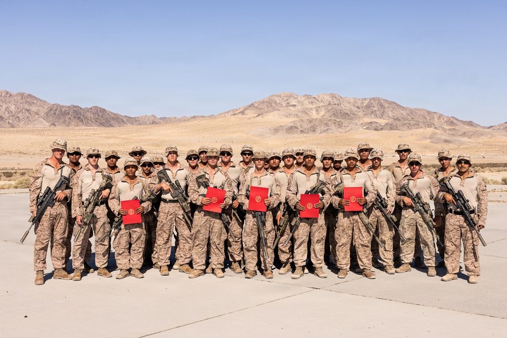 A group of approximately 25 military personnel in desert camouflage uniforms pose for a formal group photograph in an arid desert environment with mountains in the background. Several members are holding red folders or certificates, suggesting a recognition or award ceremony. The personnel are armed with rifles and wearing MARPAT (Marine pattern) desert camouflage, consistent with U.S. Marine Corps uniforms.