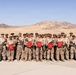 A group of approximately 25 military personnel in desert camouflage uniforms pose for a formal group photograph in an arid desert environment with mountains in the background. Several members are holding red folders or certificates, suggesting a recognition or award ceremony. The personnel are armed with rifles and wearing MARPAT (Marine pattern) desert camouflage, consistent with U.S. Marine Corps uniforms.