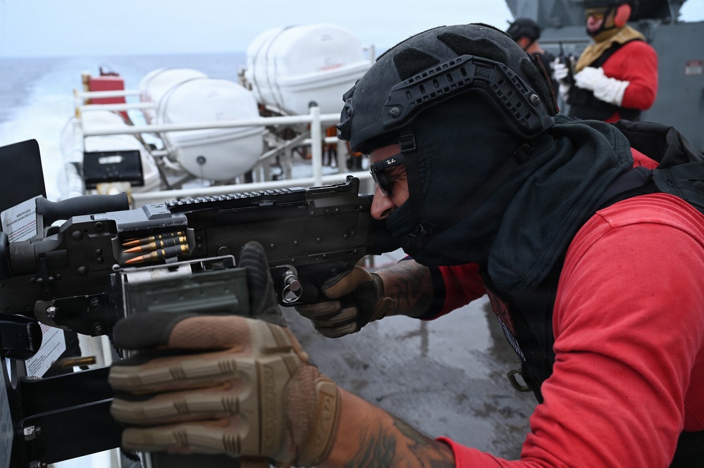 A military or naval personnel member in tactical gear is aiming a belt-fed machine gun mounted on what appears to be a naval vessel deck. The individual is wearing a black tactical helmet, balaclava, protective eyewear, body armor, and a red long-sleeve shirt with tan tactical gloves. At least one additional personnel member in similar red attire is visible in the background.