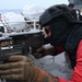 A military or naval personnel member in tactical gear is aiming a belt-fed machine gun mounted on what appears to be a naval vessel deck. The individual is wearing a black tactical helmet, balaclava, protective eyewear, body armor, and a red long-sleeve shirt with tan tactical gloves. At least one additional personnel member in similar red attire is visible in the background.