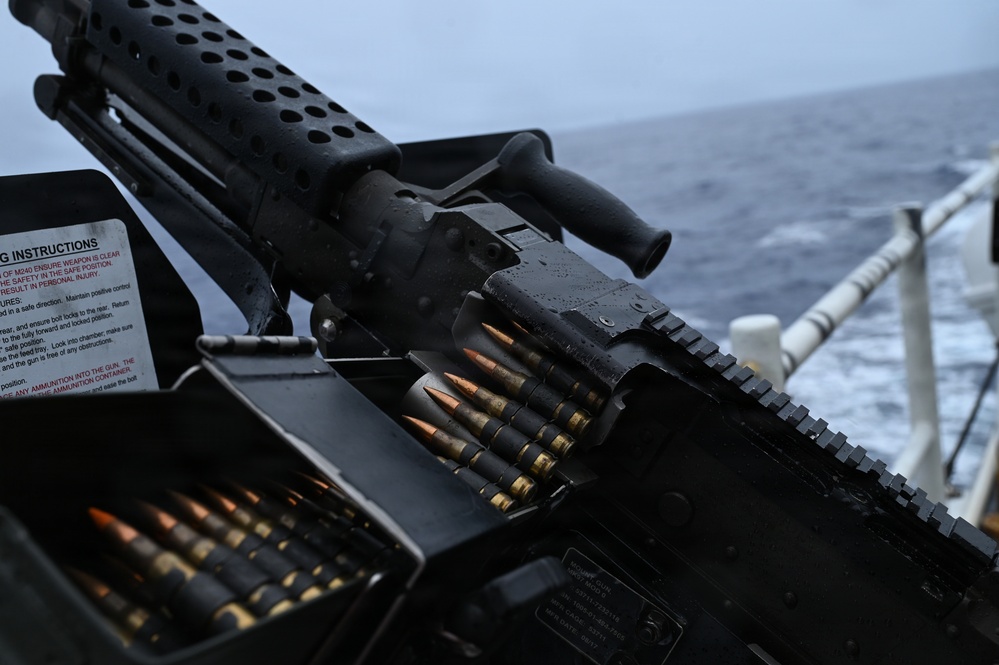 The image shows a belt-fed machine gun mounted on what appears to be a naval vessel, with a linked ammunition belt loaded and ready. A safety/operating instructions placard is visible on the left side of the mount. The weapon is positioned overlooking open ocean water, consistent with a shipboard defensive or offensive weapons station.