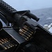 The image shows a belt-fed machine gun mounted on what appears to be a naval vessel, with a linked ammunition belt loaded and ready. A safety/operating instructions placard is visible on the left side of the mount. The weapon is positioned overlooking open ocean water, consistent with a shipboard defensive or offensive weapons station.
