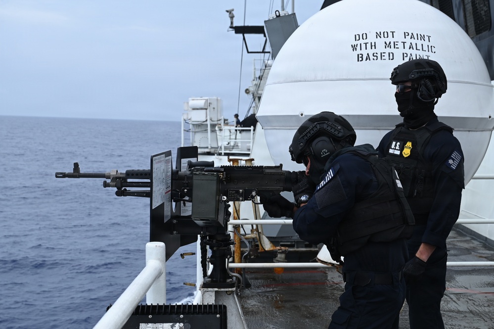 Two heavily armed personnel in black tactical gear are positioned at a mounted machine gun on the deck of a vessel at sea. The individual on the right displays a visible badge and 'U.S.' insignia on their vest, indicating a federal law enforcement affiliation. The scene appears to be a training exercise or operational readiness drill aboard a government vessel.