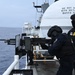 Two heavily armed personnel in black tactical gear are positioned at a mounted machine gun on the deck of a vessel at sea. The individual on the right displays a visible badge and 'U.S.' insignia on their vest, indicating a federal law enforcement affiliation. The scene appears to be a training exercise or operational readiness drill aboard a government vessel.