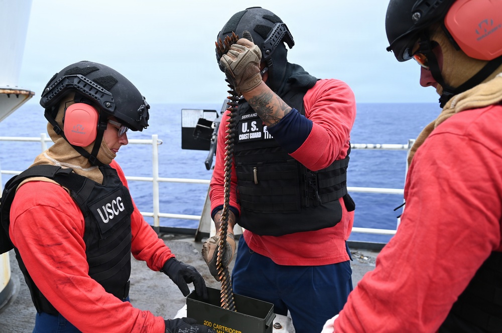 Three U.S. Coast Guard personnel are on the deck of a vessel at sea, handling a belt of ammunition. They are wearing tactical gear including helmets, body armor marked 'USCG' and 'U.S. Coast Guard,' and pink hearing protection ear muffs. The scene appears to be a weapons training or preparation exercise aboard a Coast Guard cutter.