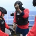 Three U.S. Coast Guard personnel are on the deck of a vessel at sea, handling a belt of ammunition. They are wearing tactical gear including helmets, body armor marked 'USCG' and 'U.S. Coast Guard,' and pink hearing protection ear muffs. The scene appears to be a weapons training or preparation exercise aboard a Coast Guard cutter.