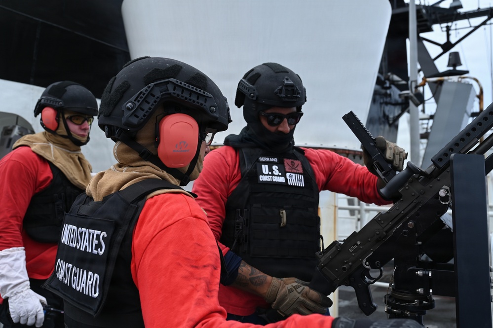 Three United States Coast Guard personnel are on the deck of a vessel, wearing tactical gear including black helmets, body armor, and red long-sleeve shirts. They appear to be conducting weapons training or maintenance on a mounted automatic weapon system. One personnel's name tape reads 'SERRANO' and is visible on their body armor.