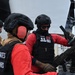 Three United States Coast Guard personnel are on the deck of a vessel, wearing tactical gear including black helmets, body armor, and red long-sleeve shirts. They appear to be conducting weapons training or maintenance on a mounted automatic weapon system. One personnel's name tape reads 'SERRANO' and is visible on their body armor.