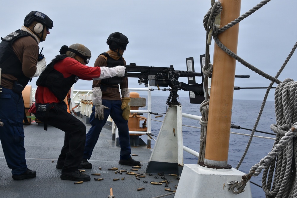 Three personnel in tactical gear and helmets are operating a mounted machine gun on the deck of a naval or coast guard vessel at sea. Spent brass casings are scattered across the deck, indicating recent or ongoing live-fire exercise. The personnel are wearing body armor, hearing protection, and gloves consistent with a weapons qualification or training drill.