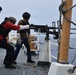 Three personnel in tactical gear and helmets are operating a mounted machine gun on the deck of a naval or coast guard vessel at sea. Spent brass casings are scattered across the deck, indicating recent or ongoing live-fire exercise. The personnel are wearing body armor, hearing protection, and gloves consistent with a weapons qualification or training drill.