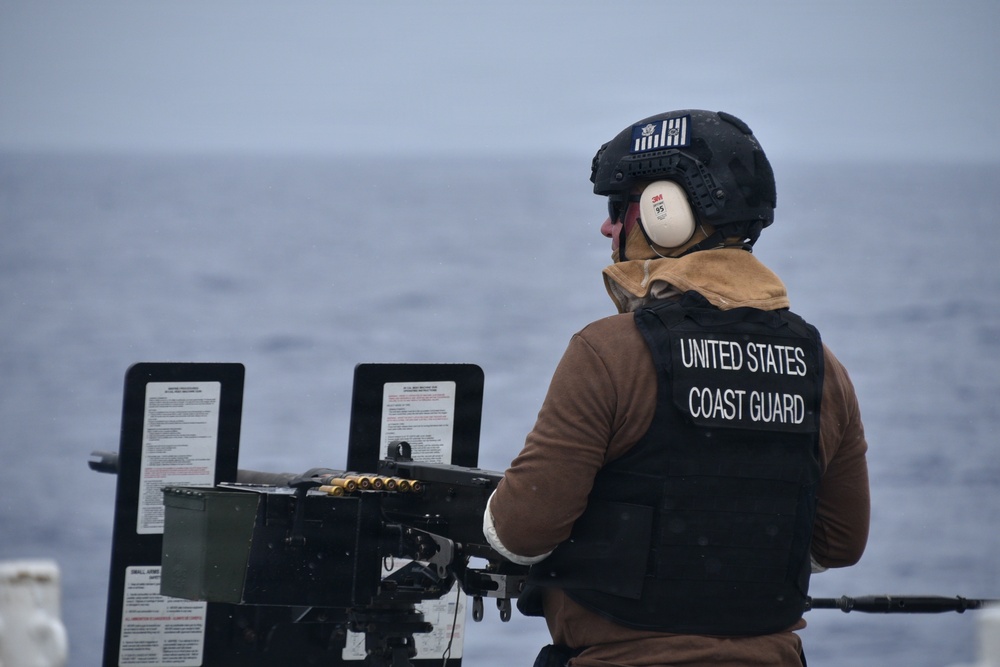 A United States Coast Guard member is positioned at a mounted weapon system aboard a vessel at sea. The personnel is wearing full protective gear including a tactical helmet, hearing protection, and a body armor vest with 'UNITED STATES COAST GUARD' printed on the back. The scene appears to be a live-fire exercise or weapons readiness drill on open water under overcast conditions.