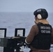 A United States Coast Guard member is positioned at a mounted weapon system aboard a vessel at sea. The personnel is wearing full protective gear including a tactical helmet, hearing protection, and a body armor vest with 'UNITED STATES COAST GUARD' printed on the back. The scene appears to be a live-fire exercise or weapons readiness drill on open water under overcast conditions.