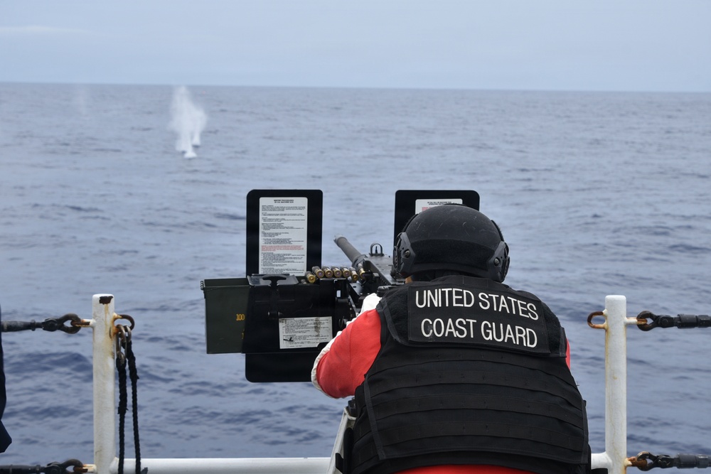 A United States Coast Guard member is positioned at a mounted machine gun on the deck of a vessel, firing at a target over open ocean. A water splash or smoke plume is visible in the distance, consistent with an active firing exercise. The scene appears to be a live-fire training or qualification exercise conducted at sea.