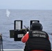A United States Coast Guard member is positioned at a mounted machine gun on the deck of a vessel, firing at a target over open ocean. A water splash or smoke plume is visible in the distance, consistent with an active firing exercise. The scene appears to be a live-fire training or qualification exercise conducted at sea.