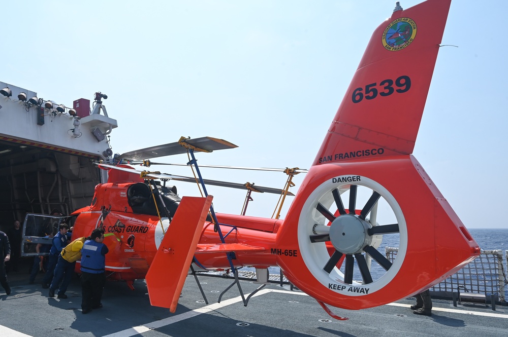 A U.S. Coast Guard MH-65E Dolphin helicopter (tail number 6539) based in San Francisco is parked on the deck of a vessel. The bright orange helicopter is secured with straps and its rotor blades are folded back. Several personnel are visible near the aircraft, appearing to conduct maintenance or inspection activities.
