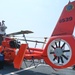 A U.S. Coast Guard MH-65E Dolphin helicopter (tail number 6539) based in San Francisco is parked on the deck of a vessel. The bright orange helicopter is secured with straps and its rotor blades are folded back. Several personnel are visible near the aircraft, appearing to conduct maintenance or inspection activities.
