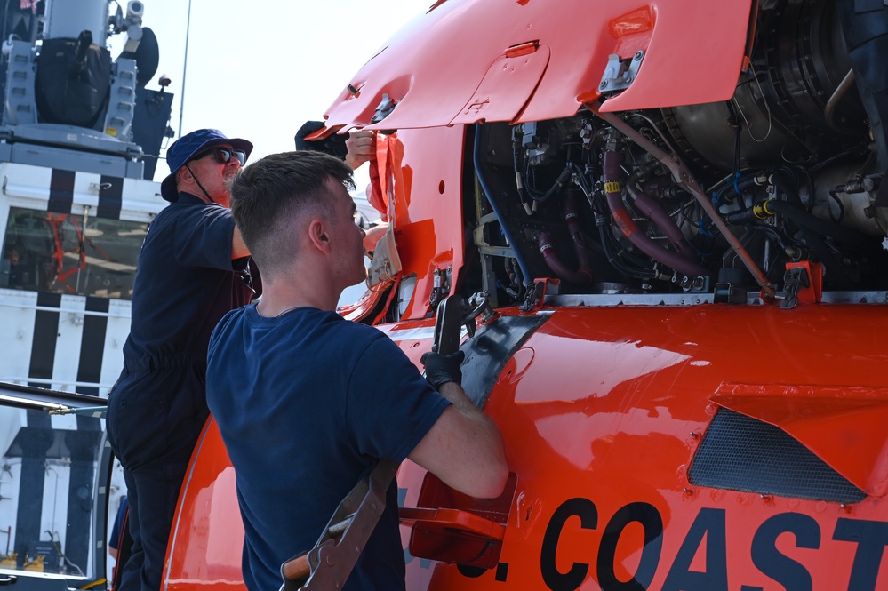 Two technicians are performing maintenance on a U.S. Coast Guard helicopter, with the engine compartment panels open and exposed. The orange aircraft bears partial text reading 'U.S. COAST' on its fuselage, consistent with a Coast Guard rotary-wing aircraft. The work appears to be taking place on the deck of a vessel or shore facility.