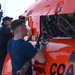 Two technicians are performing maintenance on a U.S. Coast Guard helicopter, with the engine compartment panels open and exposed. The orange aircraft bears partial text reading 'U.S. COAST' on its fuselage, consistent with a Coast Guard rotary-wing aircraft. The work appears to be taking place on the deck of a vessel or shore facility.