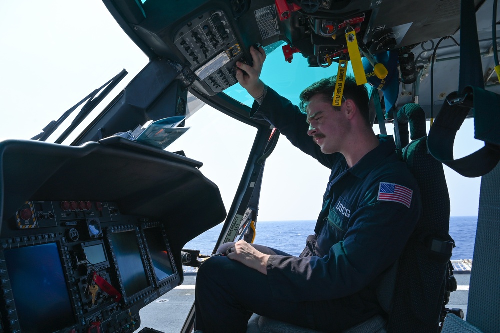 A United States Coast Guard (USCG) aviator is seated in the cockpit of a helicopter, performing a pre-flight or systems check. The individual is reaching up to interact with overhead panel controls while appearing to review documentation on their lap. The setting appears to be on a ship's flight deck over open ocean, visible through the cockpit windows.