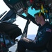 A United States Coast Guard (USCG) aviator is seated in the cockpit of a helicopter, performing a pre-flight or systems check. The individual is reaching up to interact with overhead panel controls while appearing to review documentation on their lap. The setting appears to be on a ship's flight deck over open ocean, visible through the cockpit windows.