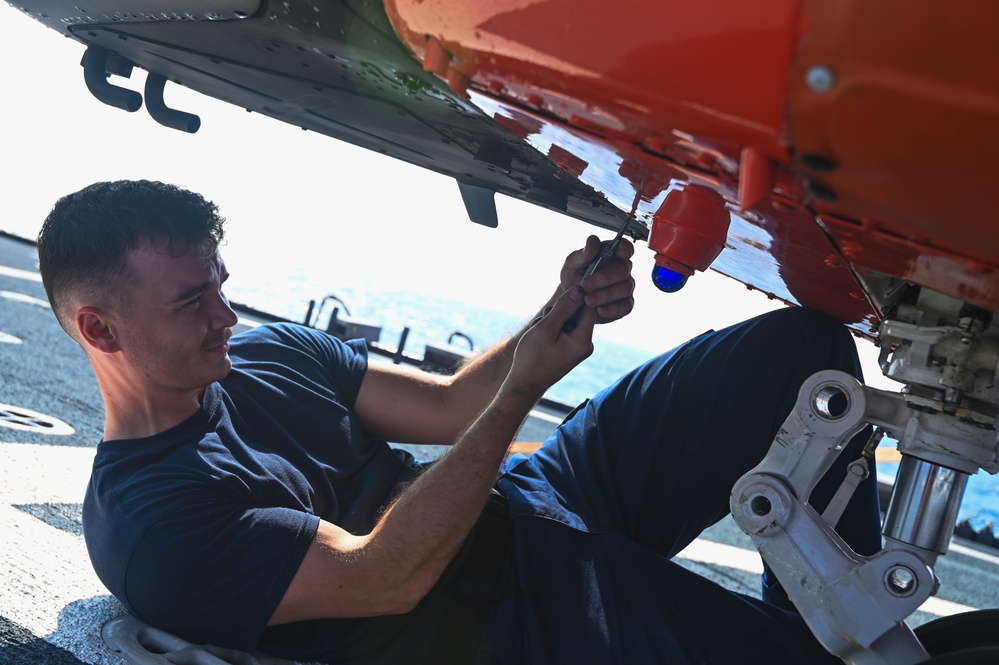 A military technician in a dark blue uniform is lying on what appears to be a flight deck, performing maintenance work on the underside of an aircraft. The aircraft features a distinctive bright orange component, likely a target drone or training missile, with a blue sensor or lens visible. The setting appears to be aboard a naval vessel at sea, based on the visible deck surface and ocean in the background.