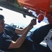 A military technician in a dark blue uniform is lying on what appears to be a flight deck, performing maintenance work on the underside of an aircraft. The aircraft features a distinctive bright orange component, likely a target drone or training missile, with a blue sensor or lens visible. The setting appears to be aboard a naval vessel at sea, based on the visible deck surface and ocean in the background.