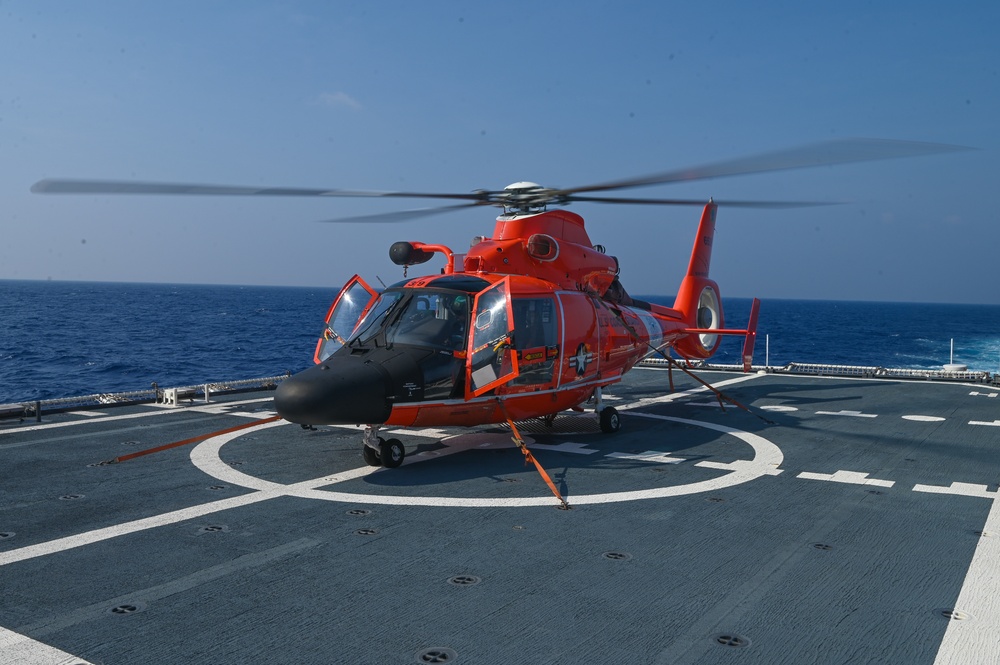 A bright orange U.S. Coast Guard MH-65 Dolphin helicopter is secured on the flight deck of a Coast Guard cutter at sea. The helicopter's rotors are spinning, indicating it is either preparing for takeoff or has just landed. The vessel appears to be underway in open ocean conditions under clear skies.