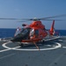 A bright orange U.S. Coast Guard MH-65 Dolphin helicopter is secured on the flight deck of a Coast Guard cutter at sea. The helicopter's rotors are spinning, indicating it is either preparing for takeoff or has just landed. The vessel appears to be underway in open ocean conditions under clear skies.