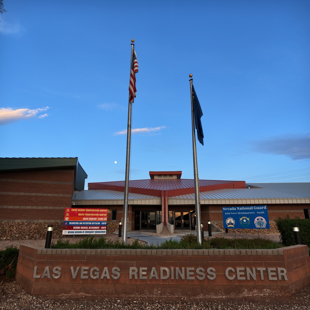 The image shows the exterior of the Las Vegas Readiness Center, a Nevada National Guard facility serving as Joint Force Headquarters South. The building features a red metal roof, brick construction, and two flagpoles flying the American flag and what appears to be the Nevada state flag. The photo was taken at dusk or dawn under a clear blue sky with a visible moon.