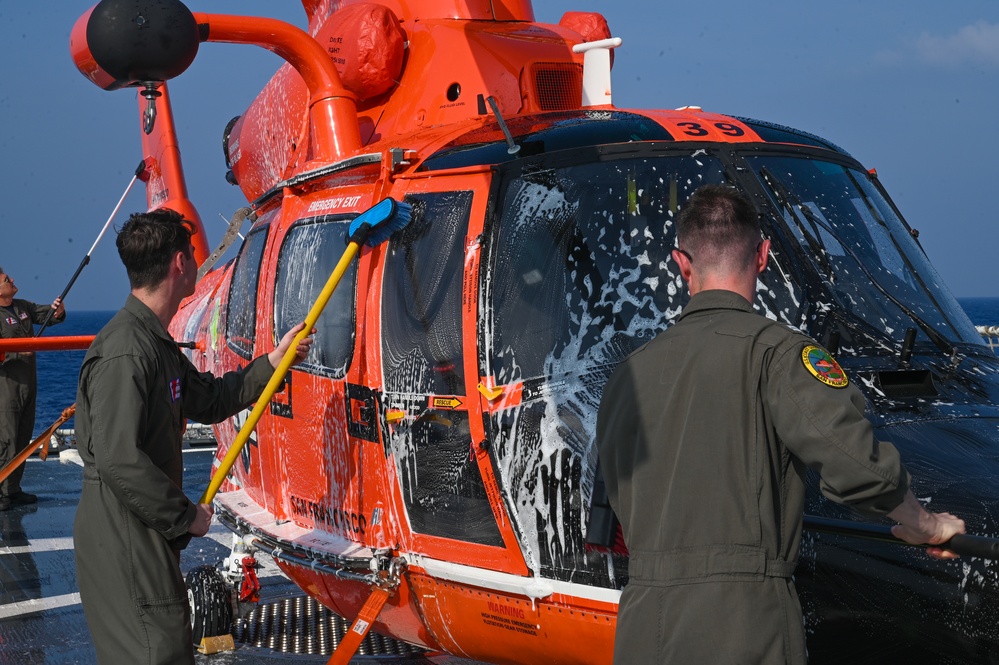 Two personnel in olive/sage green flight suits are washing an orange Coast Guard helicopter numbered '39' on what appears to be a ship's flight deck. The helicopter is covered in soap suds and is being scrubbed with long-handled brushes and sponges. The ocean is visible in the background, confirming an at-sea location.
