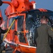 Two personnel in olive/sage green flight suits are washing an orange Coast Guard helicopter numbered '39' on what appears to be a ship's flight deck. The helicopter is covered in soap suds and is being scrubbed with long-handled brushes and sponges. The ocean is visible in the background, confirming an at-sea location.