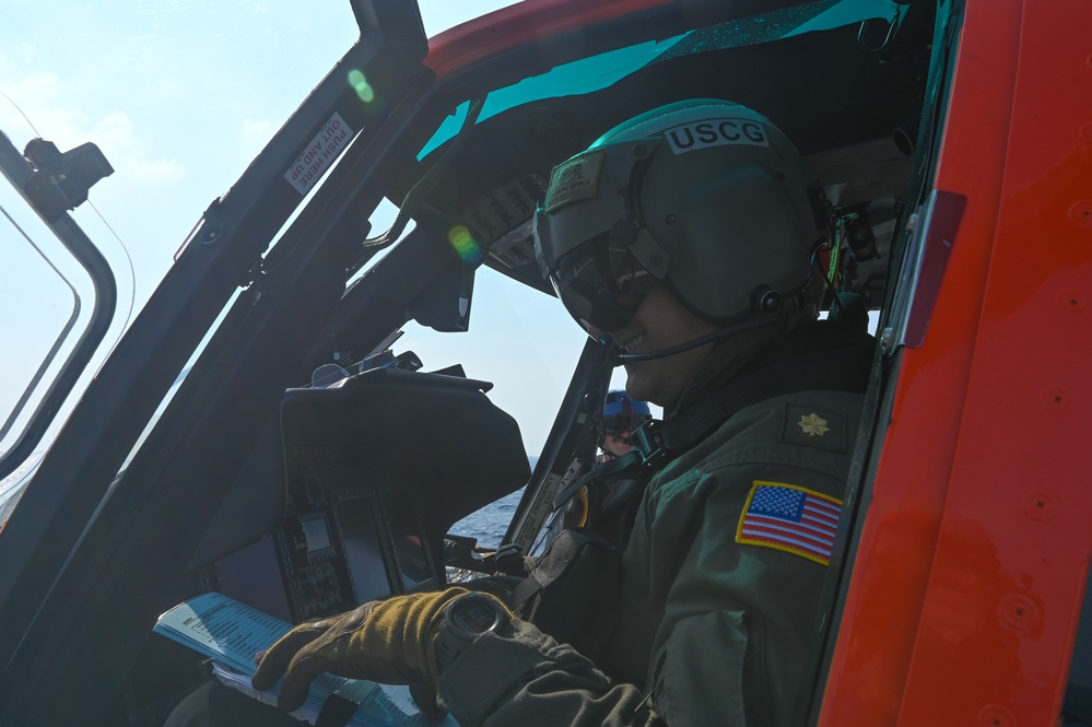 A United States Coast Guard (USCG) aviator is seated in the cockpit of a helicopter, identifiable by the 'USCG' marking on the flight helmet and the American flag patch on the flight suit. The pilot appears to be reviewing documents or a checklist while wearing full flight gear including a helmet with visor, flight gloves, and a green flight suit. A second crew member is partially visible in the background through the cockpit.
