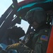 A United States Coast Guard (USCG) aviator is seated in the cockpit of a helicopter, identifiable by the 'USCG' marking on the flight helmet and the American flag patch on the flight suit. The pilot appears to be reviewing documents or a checklist while wearing full flight gear including a helmet with visor, flight gloves, and a green flight suit. A second crew member is partially visible in the background through the cockpit.