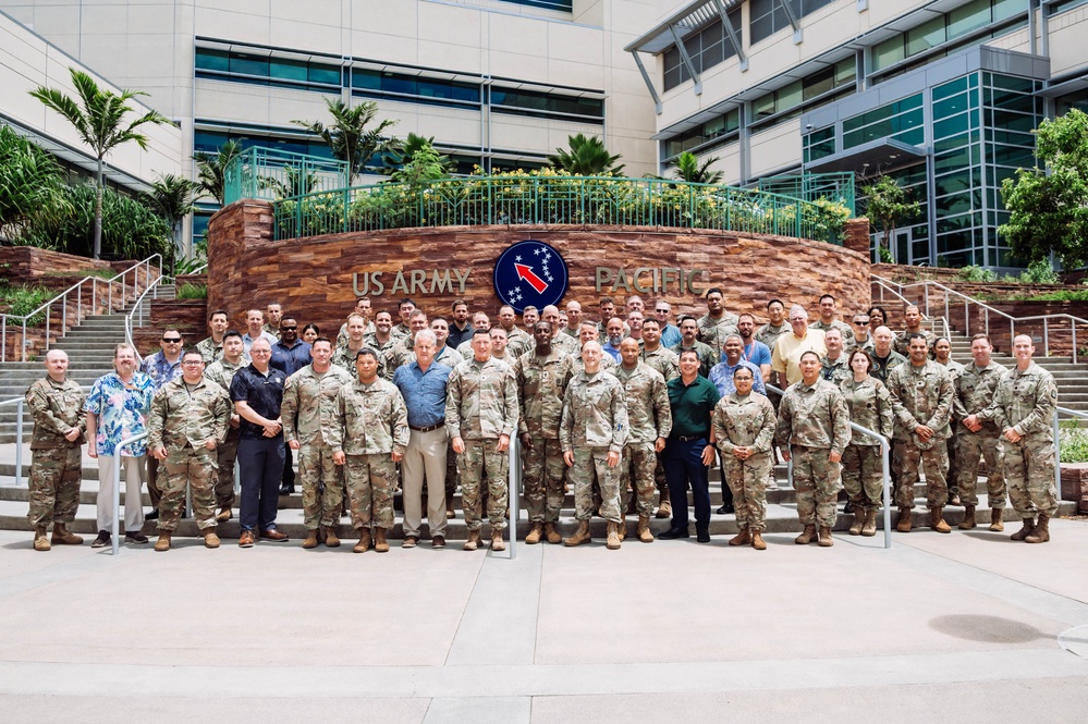 A large group of approximately 50-60 military personnel and civilians pose for a formal group photograph in front of the US Army Pacific headquarters building. The group is arranged in two rows on a plaza area, with the distinctive US Army Pacific insignia — featuring a red arrow and stars on a blue circular emblem — prominently displayed on a curved stone wall behind them. The setting appears to be in Hawaii, consistent with US Army Pacific (USARPAC) headquarters at Fort Shafter.