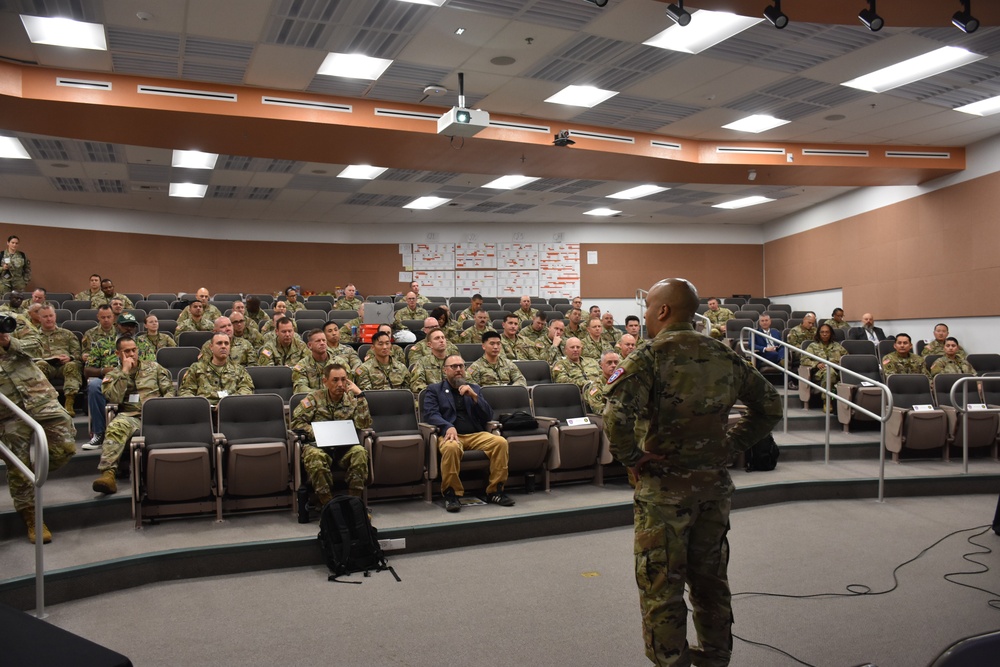 A military briefing or training session is taking place in a tiered auditorium-style room. A single uniformed soldier stands at the front addressing a large audience of approximately 50-70 military personnel, most wearing camouflage uniforms. A small number of civilians in plain clothes are also present among the audience.