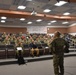 A military briefing or training session is taking place in a tiered auditorium-style room. A single uniformed soldier stands at the front addressing a large audience of approximately 50-70 military personnel, most wearing camouflage uniforms. A small number of civilians in plain clothes are also present among the audience.