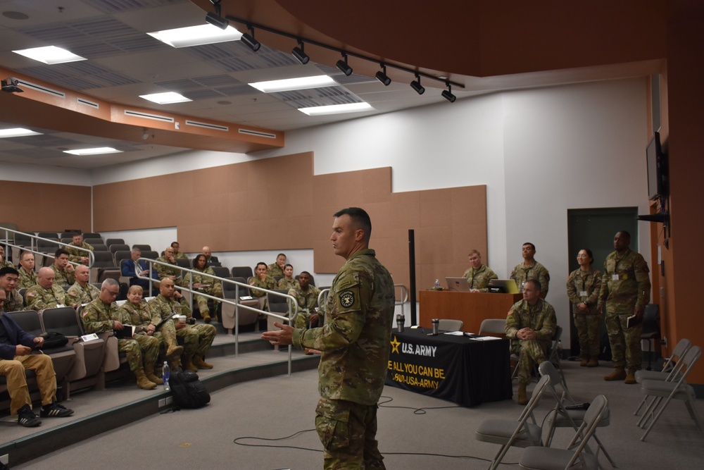 A U.S. Army soldier is addressing a group of military personnel in a tiered auditorium or briefing room. A U.S. Army branded table with the slogan 'BE ALL YOU CAN BE' and '1-800-USA-ARMY' is visible in the background. The audience consists of uniformed soldiers seated in stadium-style seating, with additional personnel standing along the walls.