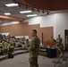 A U.S. Army soldier is addressing a group of military personnel in a tiered auditorium or briefing room. A U.S. Army branded table with the slogan 'BE ALL YOU CAN BE' and '1-800-USA-ARMY' is visible in the background. The audience consists of uniformed soldiers seated in stadium-style seating, with additional personnel standing along the walls.