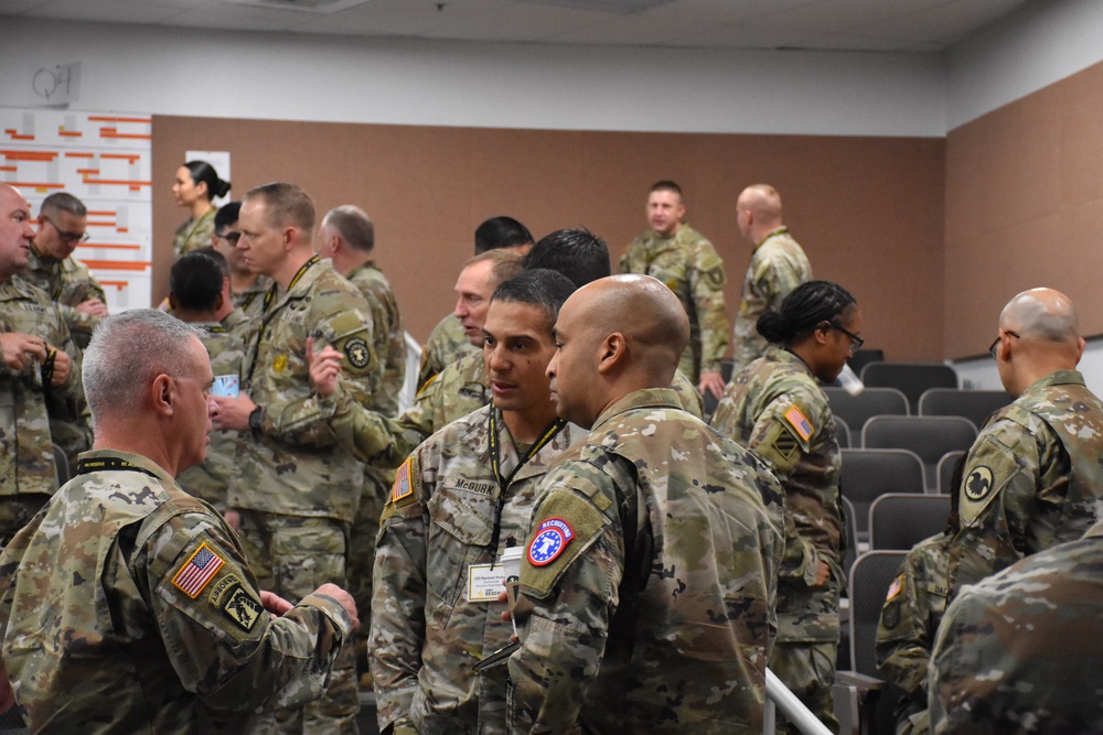 A group of U.S. Army soldiers in OCP (Operational Camouflage Pattern) uniforms are gathered in what appears to be a conference or briefing room. The soldiers are engaged in informal conversations and networking, with several wearing name tags and unit patches visible on their uniforms. The setting suggests a military training event, conference, or professional development gathering.