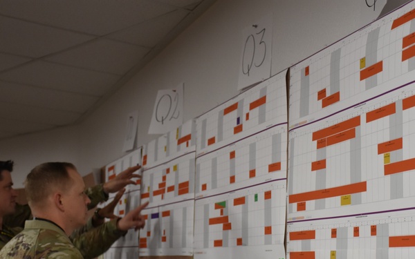 Two U.S. Army soldiers in MultiCam/OCP camouflage uniforms are reviewing large planning charts or scheduling boards mounted on a wall. The boards appear to be quarterly training or operational schedules labeled 'Q2' and 'Q3', featuring color-coded Gantt-style charts with orange, gray, and other colored bars. One soldier is pointing at specific elements on the charts while the other observes.
