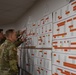 Two U.S. Army soldiers in MultiCam/OCP camouflage uniforms are reviewing large planning charts or scheduling boards mounted on a wall. The boards appear to be quarterly training or operational schedules labeled 'Q2' and 'Q3', featuring color-coded Gantt-style charts with orange, gray, and other colored bars. One soldier is pointing at specific elements on the charts while the other observes.