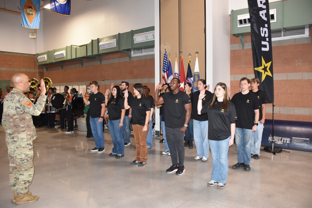 A U.S. Army enlistment ceremony is taking place in what appears to be a large indoor facility such as an armory or gymnasium. A uniformed soldier in Army Combat Uniform (ACU) is administering the oath of enlistment to a group of approximately 12-15 recruits, all wearing matching black U.S. Army t-shirts with the Army star logo. A military band and color guard with multiple flags are visible in the background.